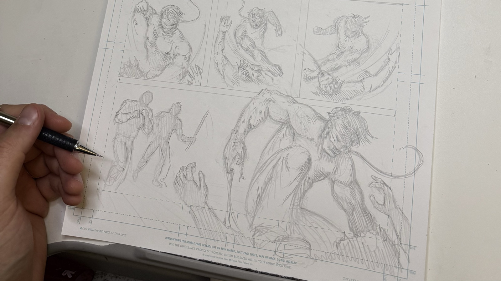 A hand pencilling a comic page on bristol board: three panels along the top and a wide action panel below